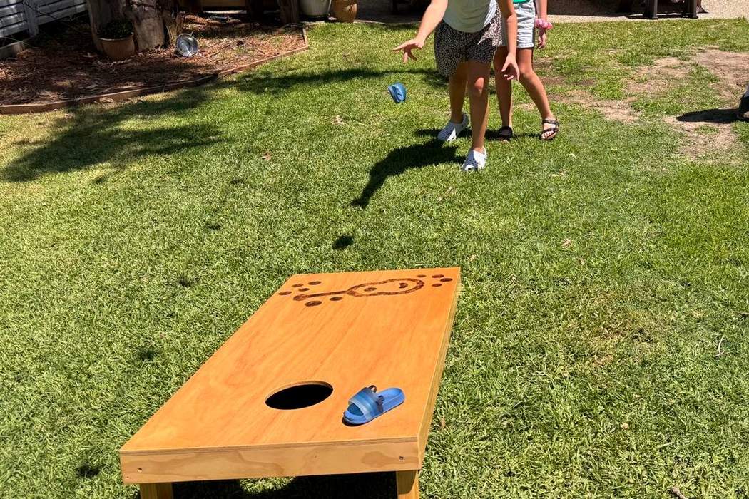 A wooden cornhole board set up on the lawn, with a guest throwing a bag