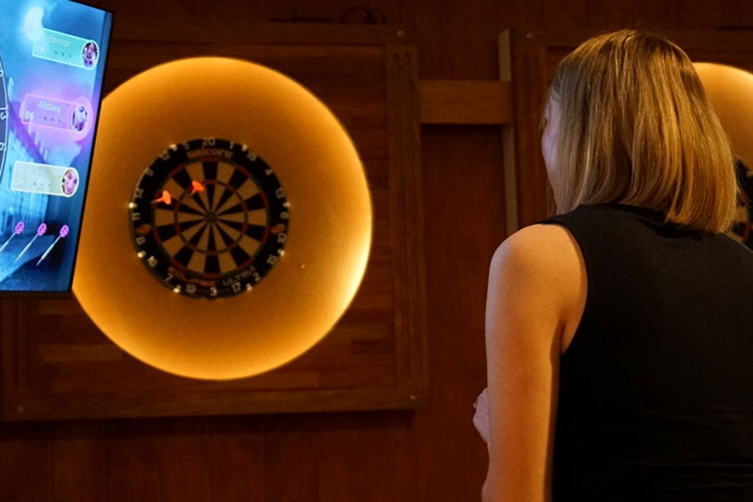 A guest playing electronic darts, with the scoring screens and lit dartboards behind her