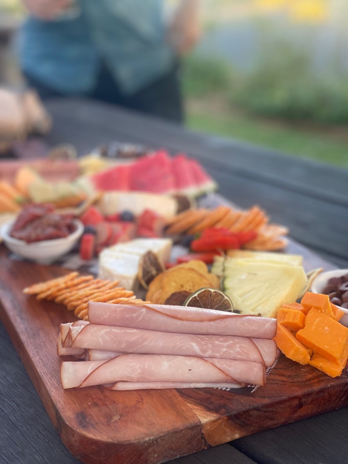A party grazing board with ham, cheeses, crackers, fruit and dips on an outdoor table
